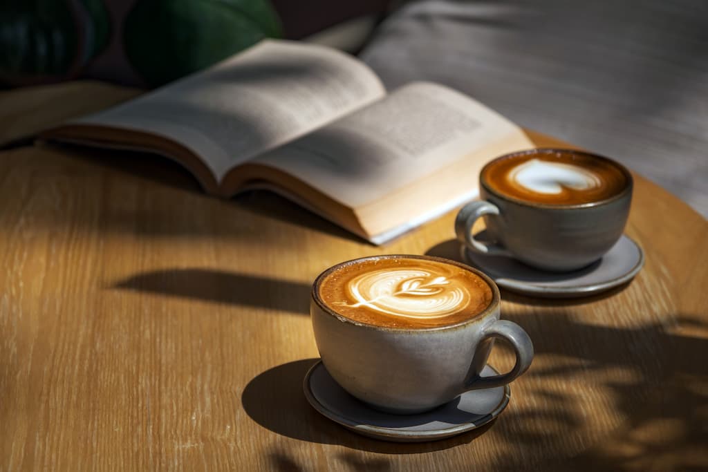 Two lattes in ceramic mugs on a wooden table next to an open book at a coffee shop near our Hershey bed and breakfast